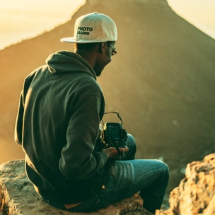 Person sitting on rocks facing right on a mountain holding a camera